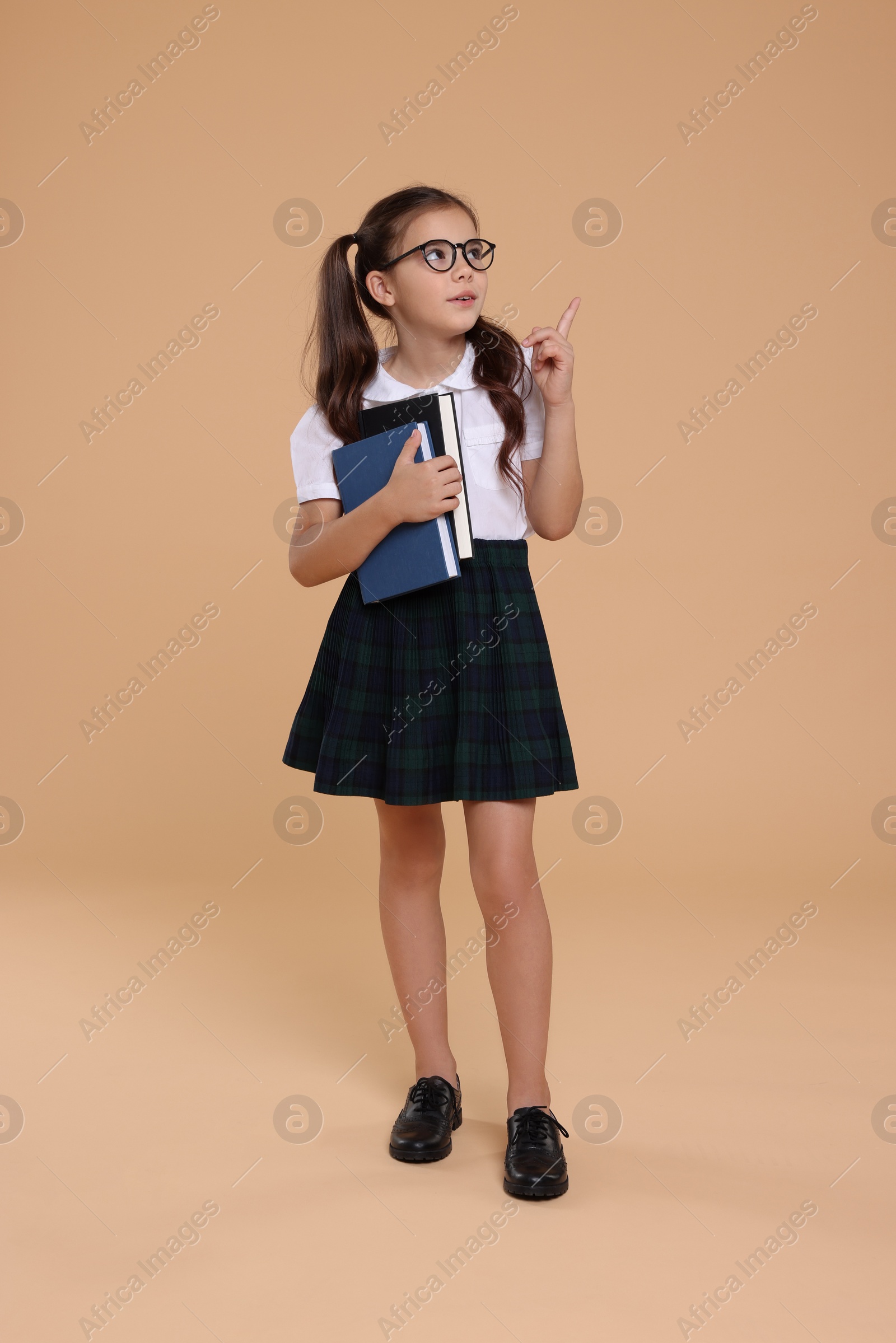 Photo of Cute schoolgirl in glasses with books pointing upwards on beige background