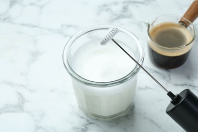 Photo of Mini mixer (milk frother), whipped milk in glass and coffee on white marble table, closeup. Space for text