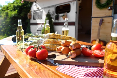 Photo of Sausages, vegetables and bottles of beer on wooden table near motorhome on sunny day. Camping season