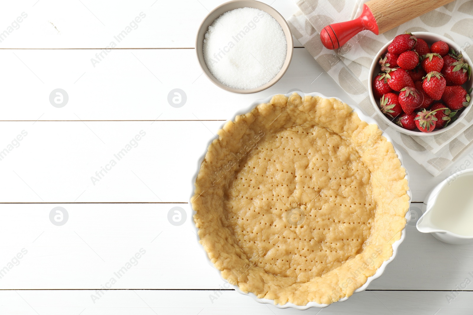Photo of Making shortcrust pastry. Raw dough in baking dish, strawberries, sugar, rolling pin and milk on white wooden table, top view. Space for text