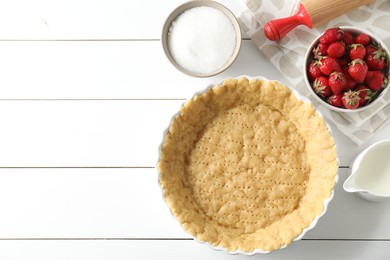 Photo of Making shortcrust pastry. Raw dough in baking dish, strawberries, sugar, rolling pin and milk on white wooden table, top view. Space for text