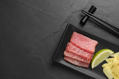 Photo of Tasty sashimi (pieces of fresh raw tuna), lime wedge and ginger slices on black table, flat lay. Space for text