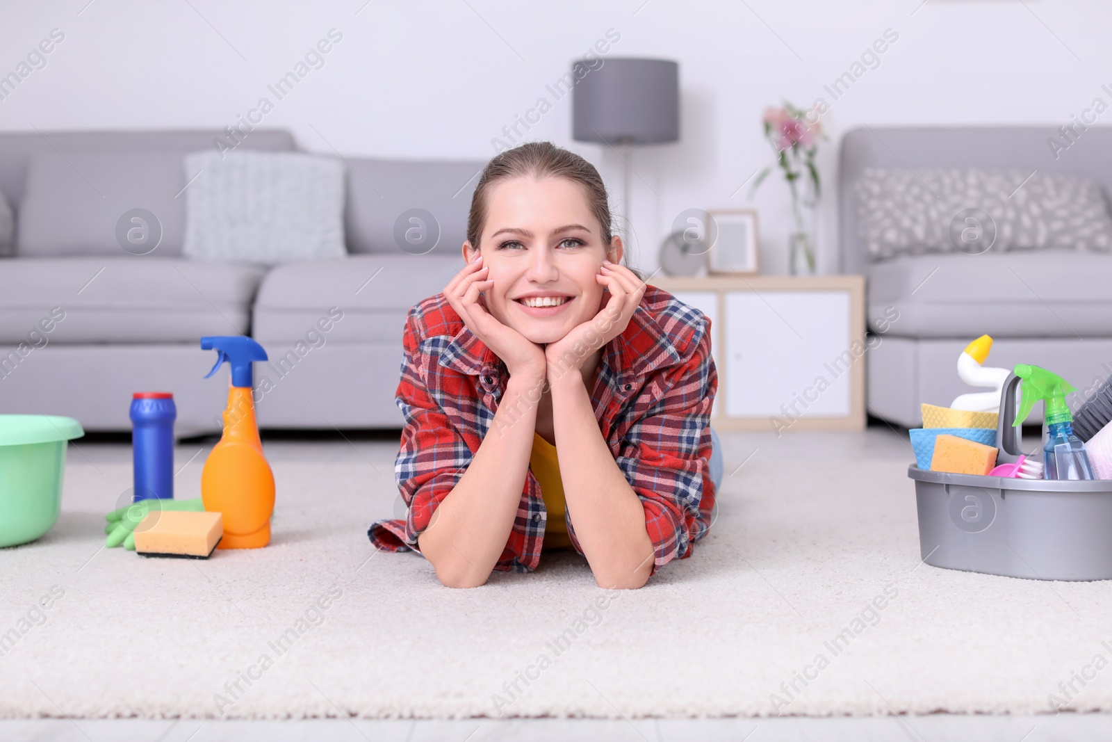 Photo of Young woman with cleaning supplies lying on carpet at home