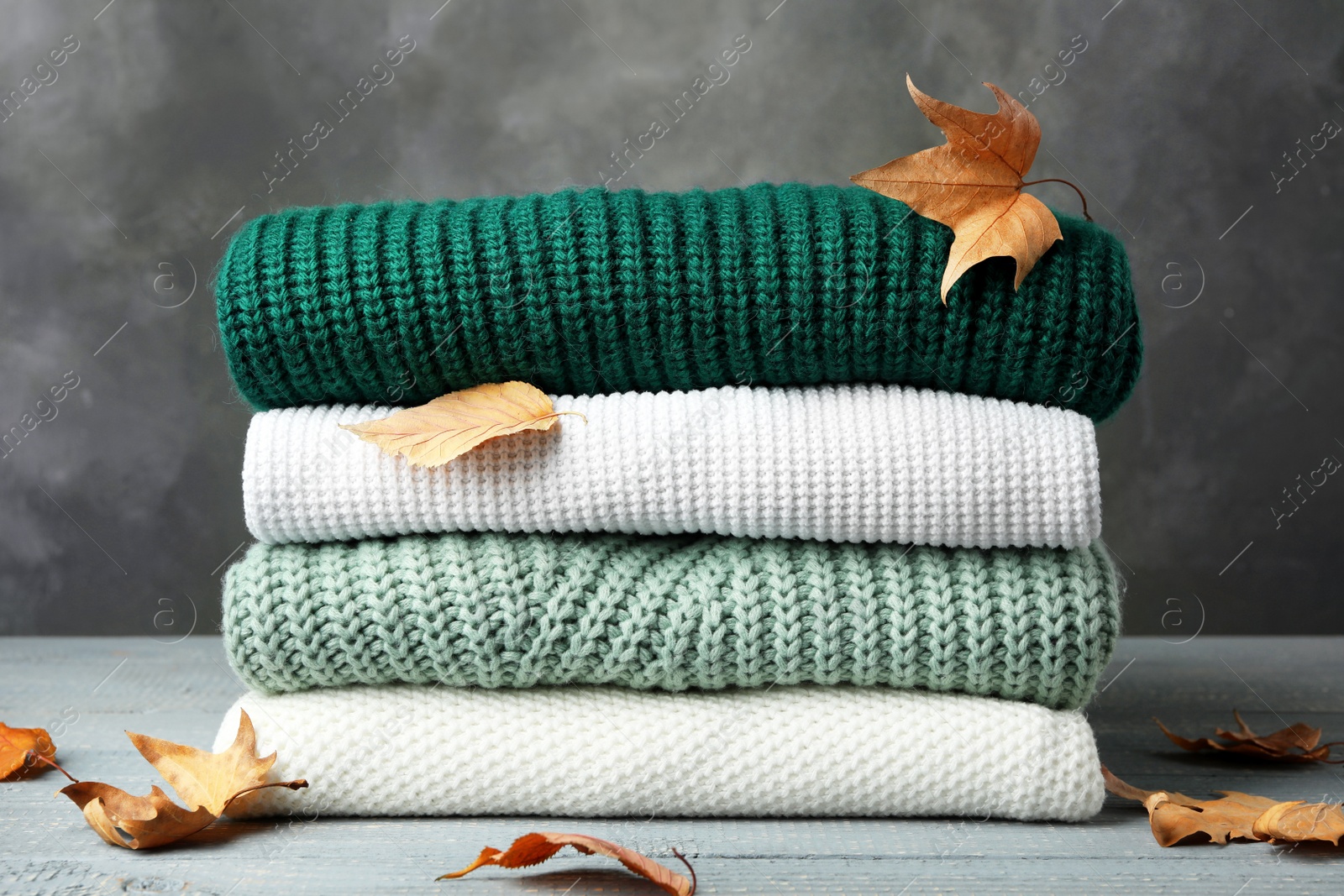 Photo of Stack of warm clothes and autumn leaves on wooden table against grey background