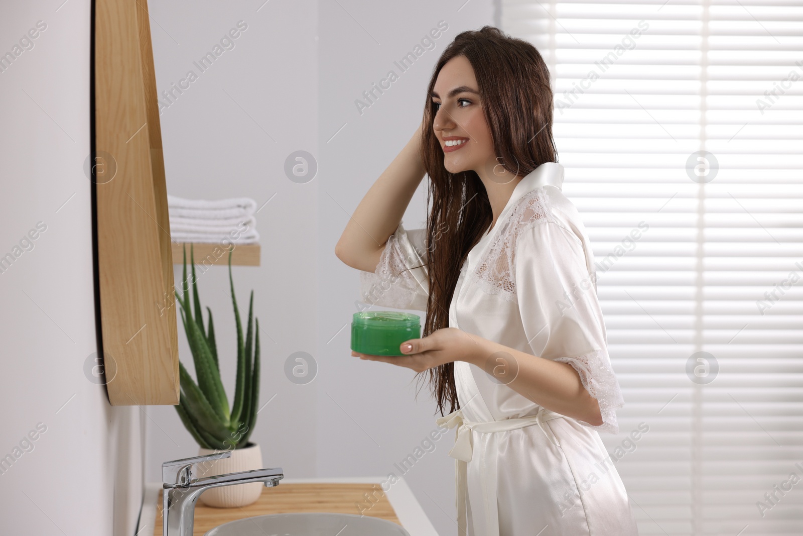 Photo of Young woman applying aloe hair mask near mirror in bathroom