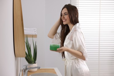 Photo of Young woman applying aloe hair mask near mirror in bathroom