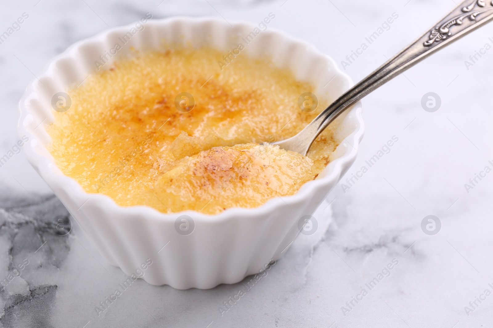 Photo of Delicious creme brulee in bowl and spoon on white marble table, closeup