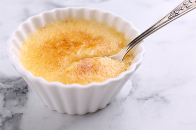 Photo of Delicious creme brulee in bowl and spoon on white marble table, closeup