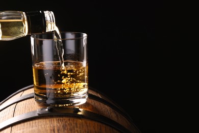 Photo of Pouring whiskey from bottle into glass on wooden barrel against dark background, closeup. Space for text