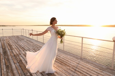 Gorgeous bride in beautiful wedding dress with bouquet near river on sunset
