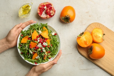 Woman holding bowl of delicious persimmon salad at light grey table, top view