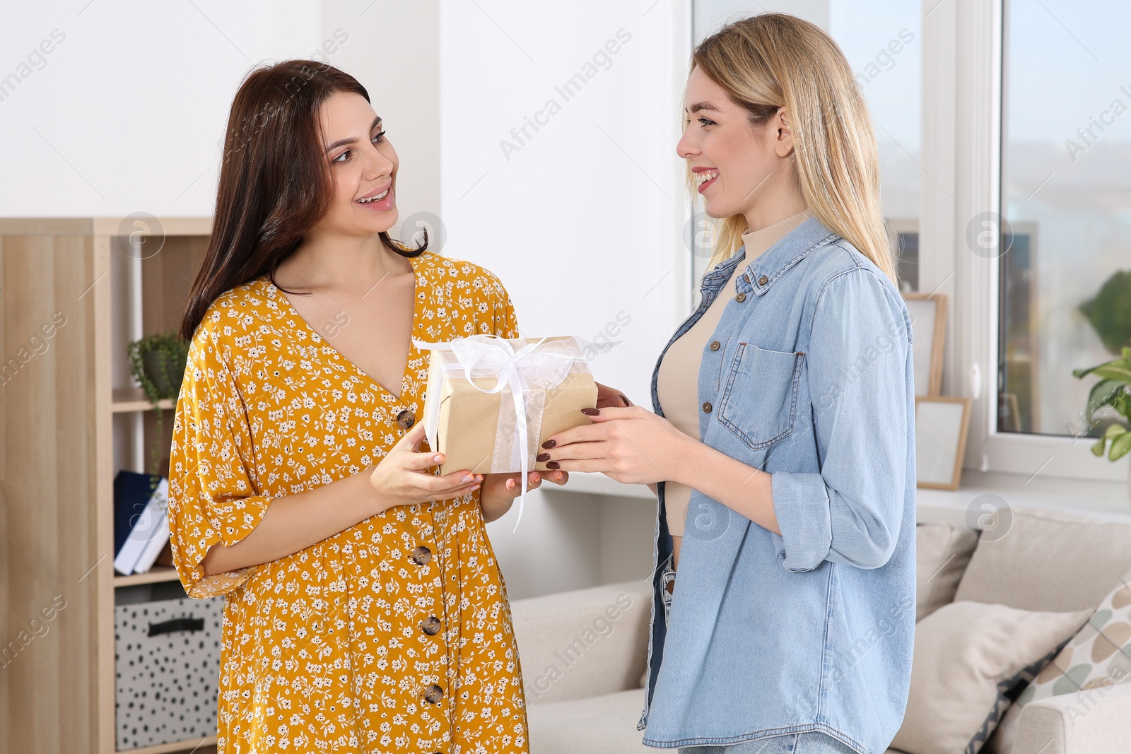 Photo of Smiling young woman presenting gift to her friend at home