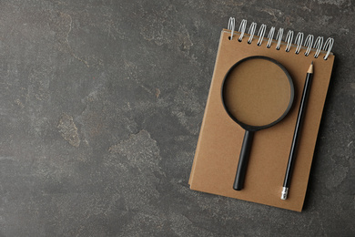 Top view of magnifier glass, empty notebook and pencil on grey stone background, space for text. Find keywords concept