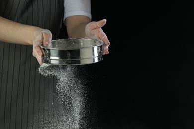 Woman sieving flour at table against black background, closeup. Space for text