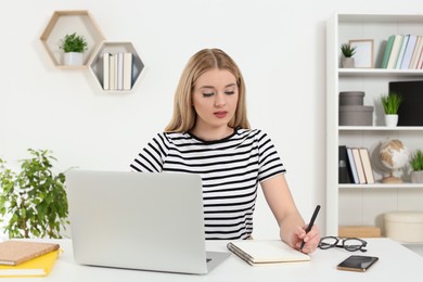 Home workplace. Woman writing in notebook near laptop at white desk in room