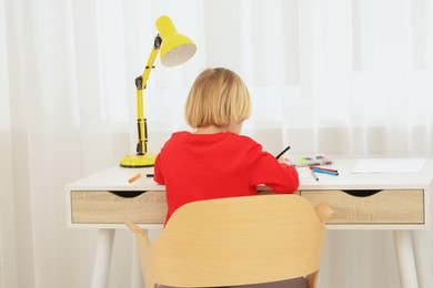 Photo of Little boy sitting at desk in room, back view. Home workplace