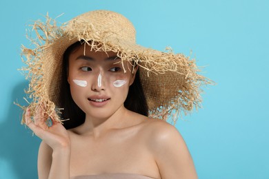 Beautiful young woman in straw hat with sun protection cream on her face against light blue background, space for text