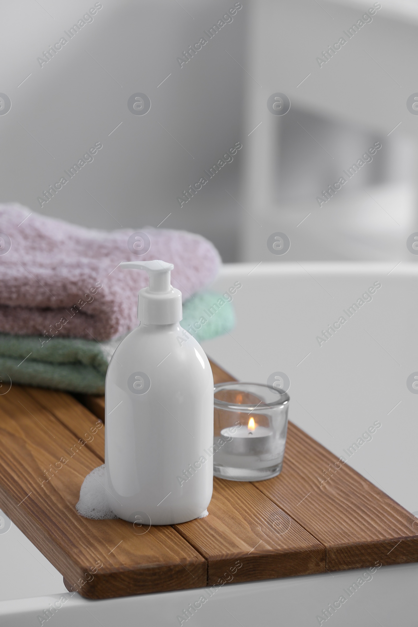 Photo of Bottle of bubble bath with foam, towels and candle on tub in bathroom