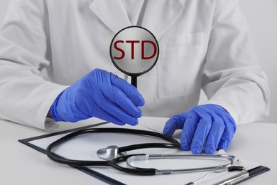 Doctor holding magnifying glass with abbreviation STD at white table, closeup