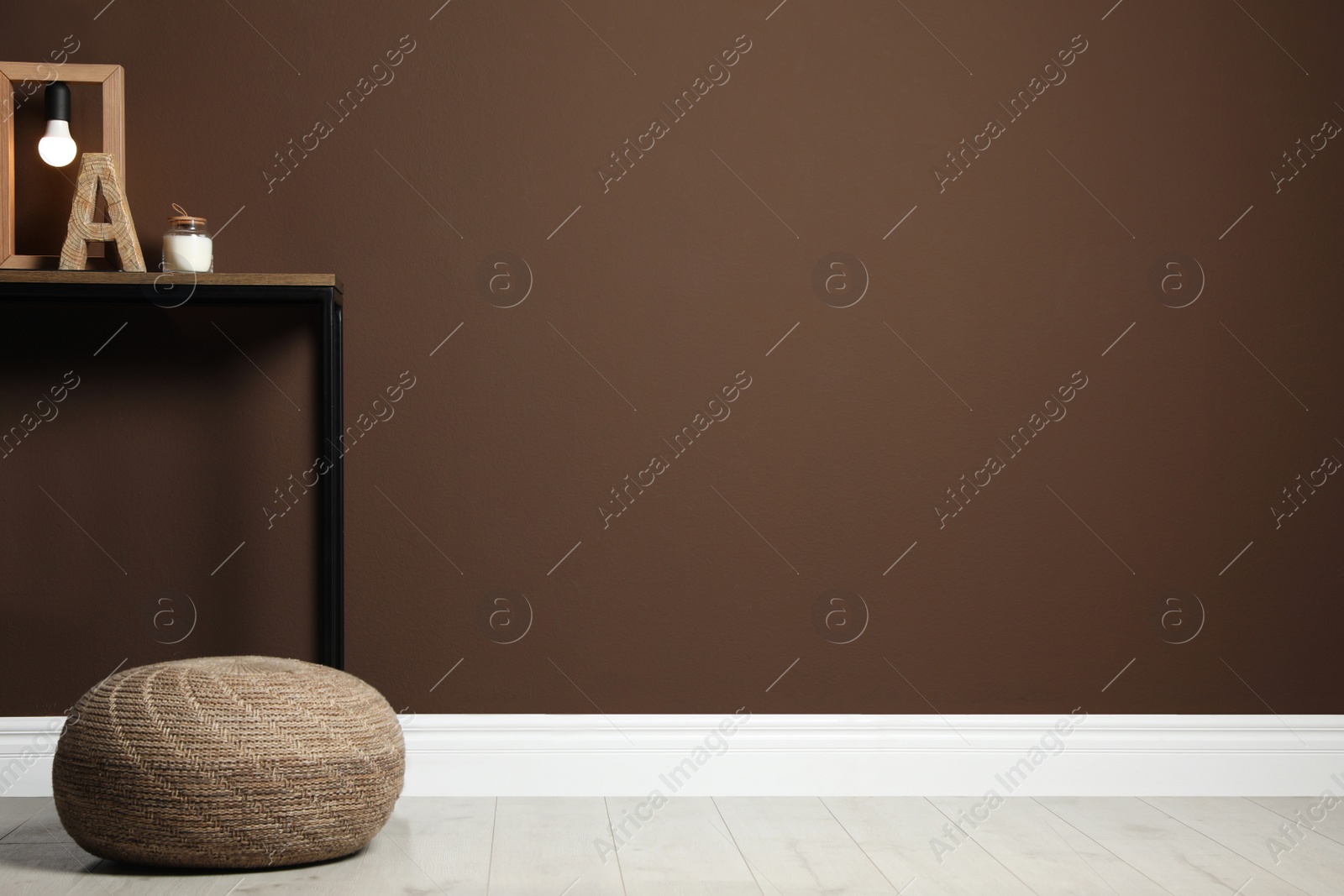 Photo of Comfortable knitted pouf and table with decor elements near brown wall indoors, space for text. Interior design