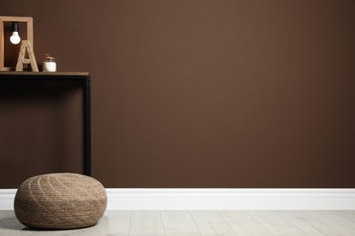 Photo of Comfortable knitted pouf and table with decor elements near brown wall indoors, space for text. Interior design