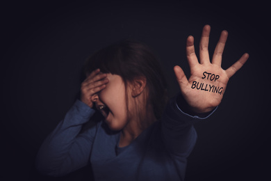 Image of Crying little girl showing palm with message STOP BULLYING near black wall, focus on hand