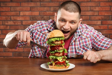 Young hungry man with cutlery and tasty huge burger at table