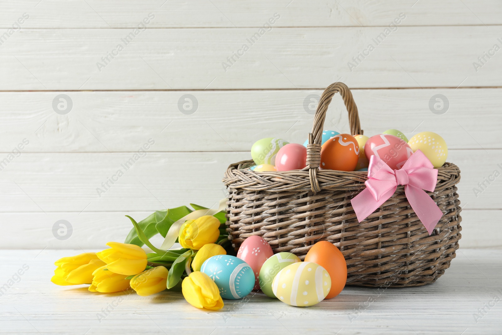 Photo of Wicker basket with painted Easter eggs and spring flowers on table, space for text