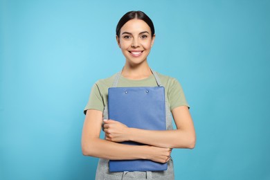Photo of Young woman in grey apron with clipboard on light blue background
