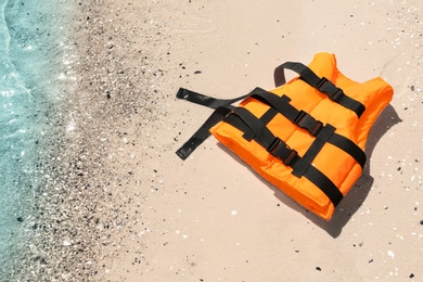 Photo of Orange life jacket on sand near sea, above view. Emergency rescue equipment