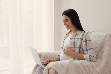 Pregnant young woman with laptop at home, space for text