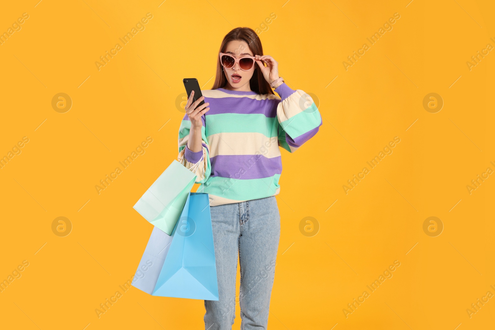 Photo of Surprised young woman with shopping bags looking at smartphone on yellow background. Big sale