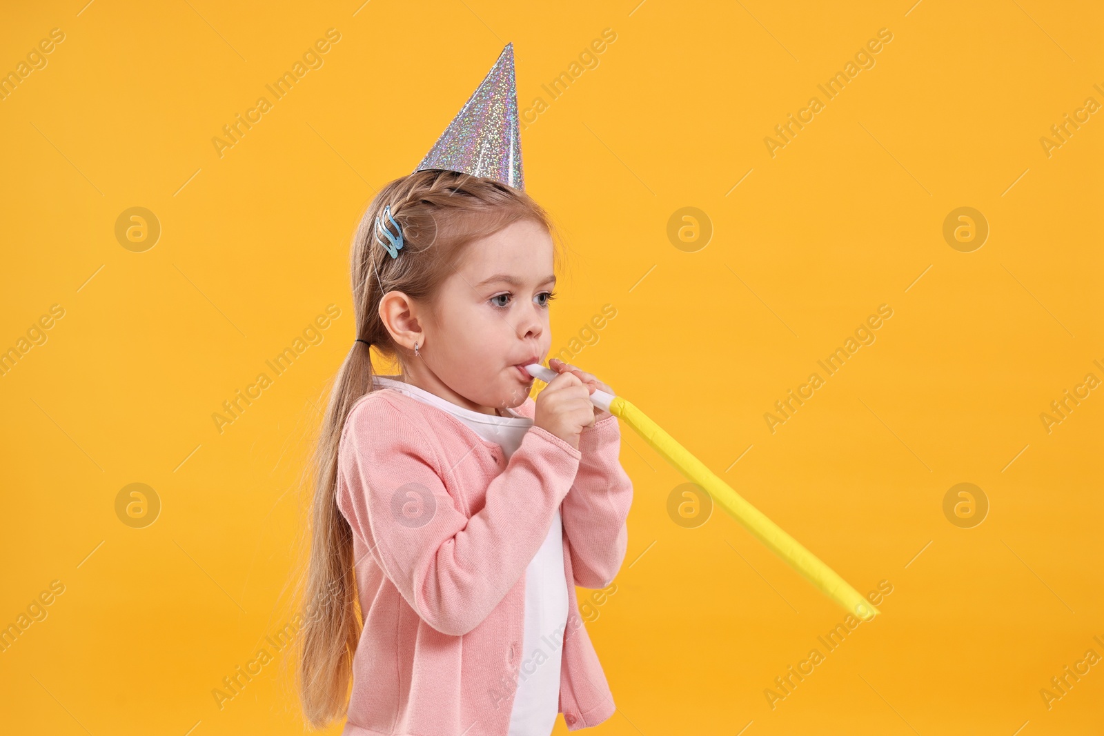 Photo of Birthday celebration. Cute little girl in party hat with blower on orange background