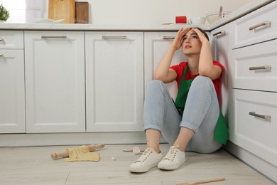 Photo of Tired woman sitting on dirty floor with utensils and food leftovers in messy kitchen