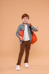 Photo of Smiling schoolboy with backpack and books showing thumb up on beige background