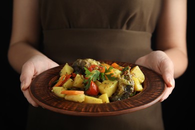 Photo of Woman holding earthenware with delicious roasted potatoes and meat on black background, closeup