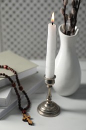 Books, burning candle, rosary beads and vase of willow branches on white table, closeup