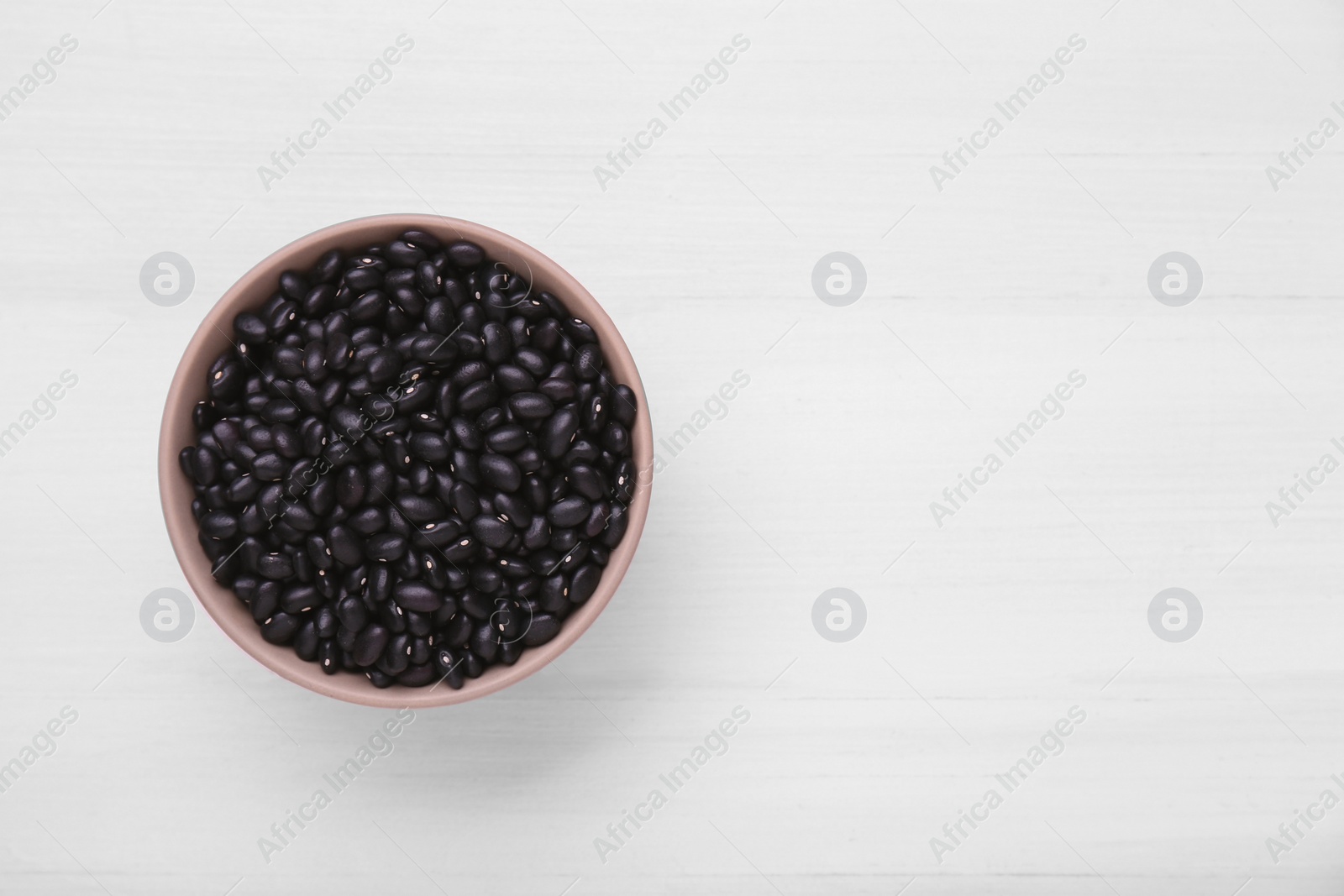 Photo of Bowl of raw black beans on white wooden table, top view. Space for text