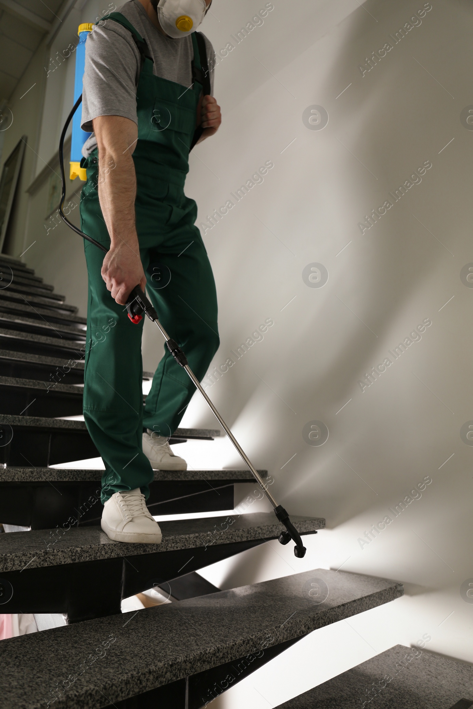 Photo of Pest control worker in uniform spraying pesticide on stairs indoors