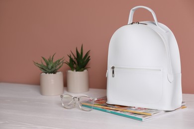 Stylish urban backpack, glasses and magazines on white wooden table