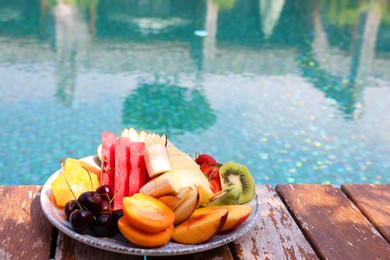 Photo of Plate with fresh fruits on wooden deck near outdoor swimming pool, space for text. Luxury resort