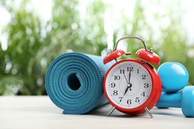 Alarm clock, dumbbells and yoga mat on table outdoors, space for text. Morning exercise