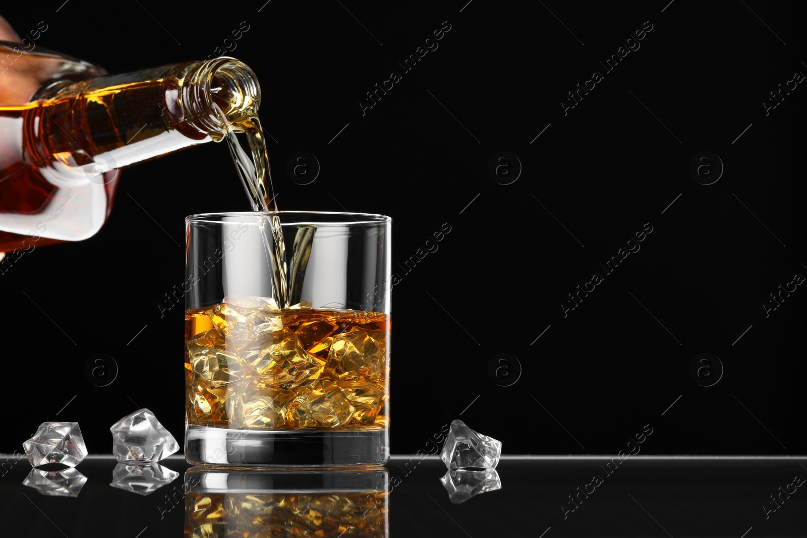 Photo of Pouring whiskey into glass with ice cubes at table against black background, space for text