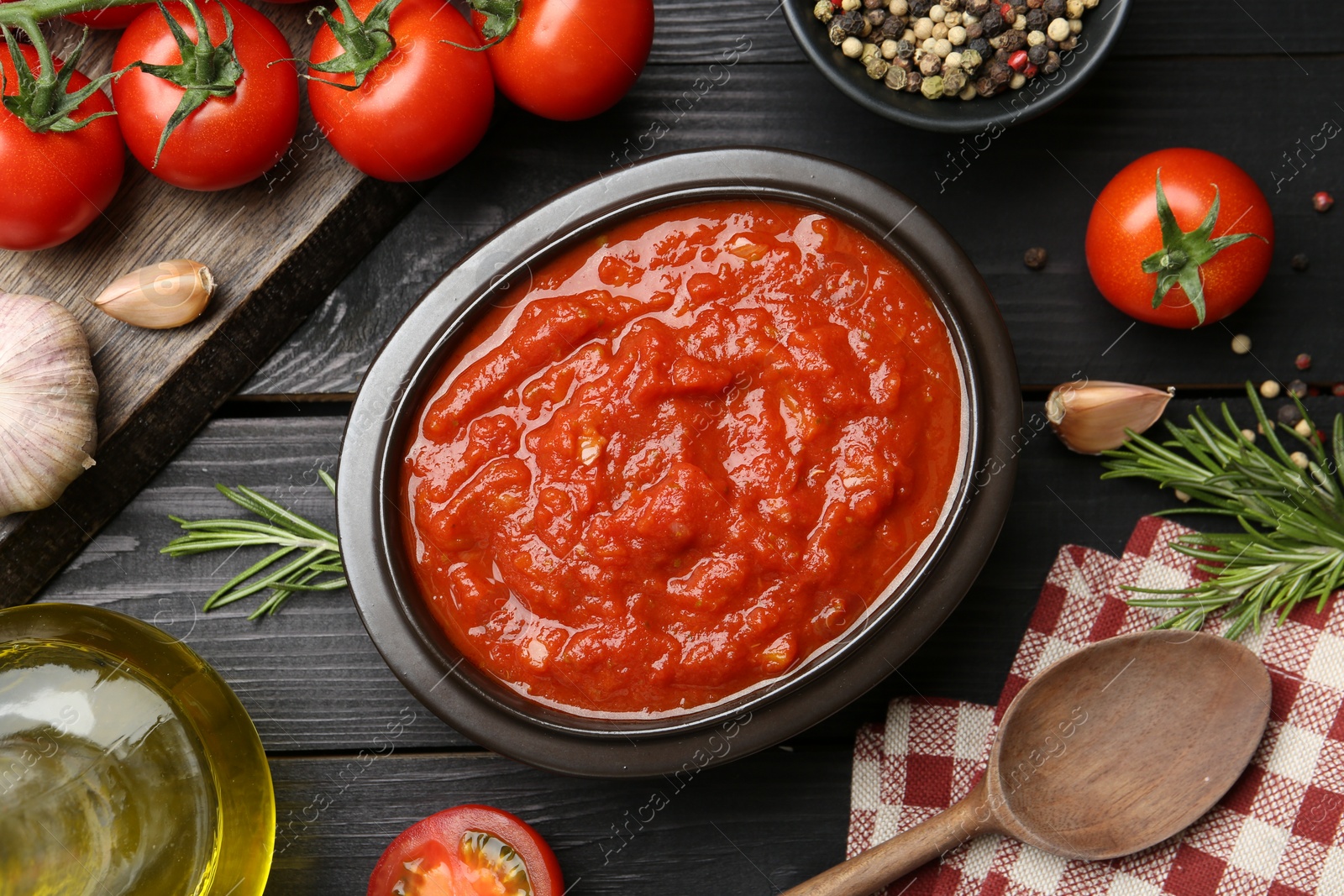 Photo of Homemade tomato sauce in bowl, spoon and fresh ingredients on black wooden table, flat lay