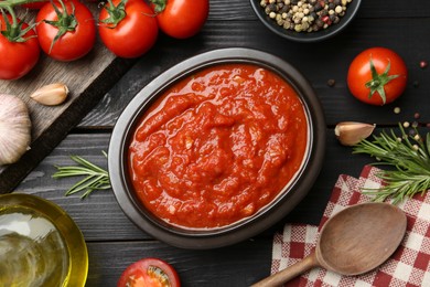 Homemade tomato sauce in bowl, spoon and fresh ingredients on black wooden table, flat lay