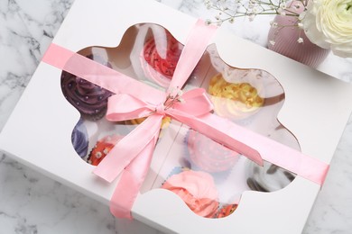 Photo of Different colorful cupcakes in box and flowers on white marble table, above view