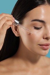 Beautiful young woman applying serum onto her face on light blue background, closeup