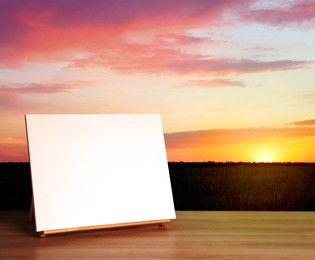 Image of Wooden easel with blank canvas on table and beautiful field at sunset. Space for text
