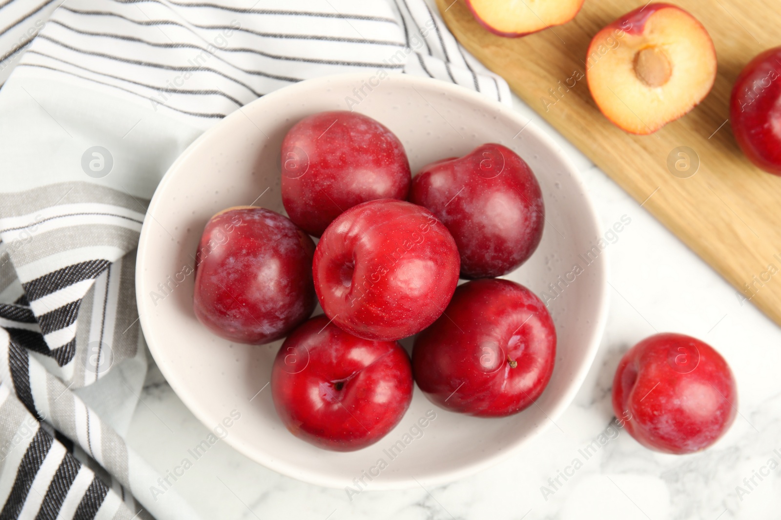 Photo of Delicious ripe plums in bowl on white marble table, flat lay