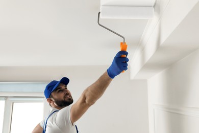 Photo of Handyman painting ceiling with white dye indoors, focus on roller